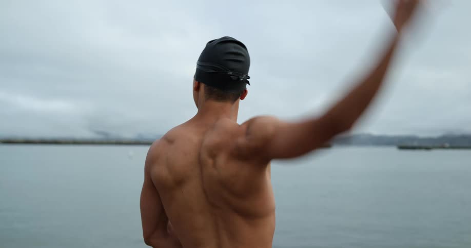 A man with goggles and a swimming cap stretching with the sky and clouds in the background