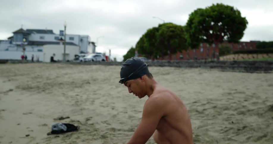 A male wearing swim trunks and a swim cap stretching on a beach