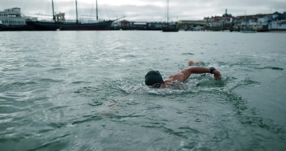 A man swims further from the shore where a marina and several buildings sit
