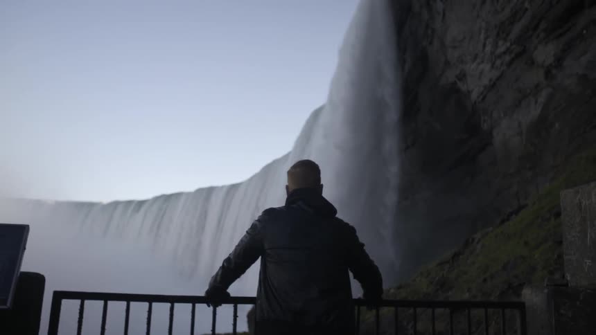 A man stands at a fence looking up at what appears to be niagara falls