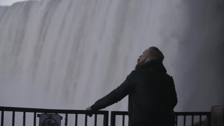 A bearded man stands near a waterfall