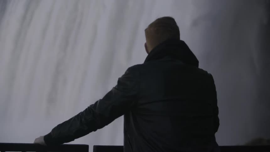 A man is leaning against a railing taking in the view of a waterfall