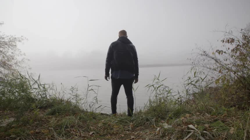 A man stands near a lake on a foggy morning
