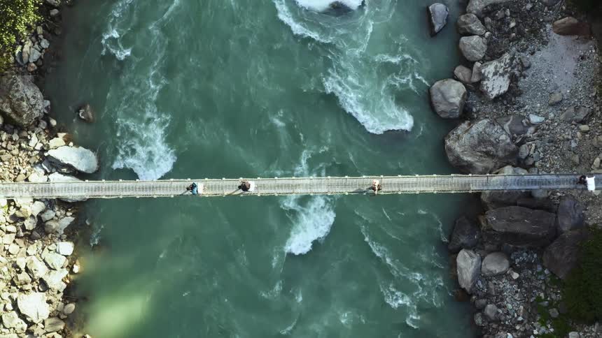 An aerial view of a small group of hikers crossing over a rushing river using a narrow walking bridge