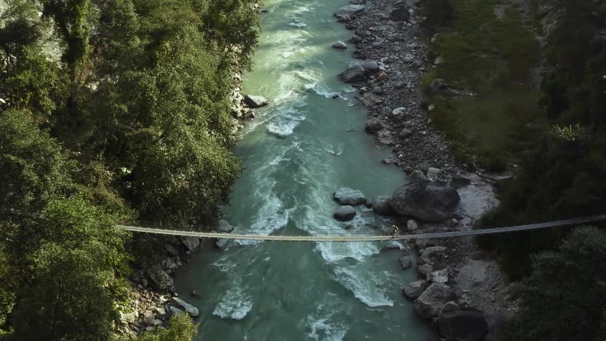 A hiker crosses a narrow suspension bridge over a river in a woodland during the day