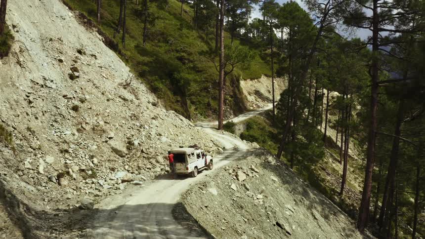Man hangs on to back of truck driving along dirt road on steep mountain slope