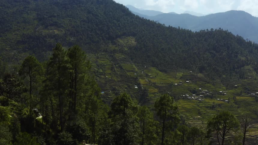 A small village next to a hill with trees with a mountain range and the sky in the background