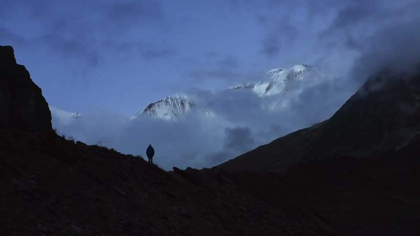Person stands in shadows mountain pass looking toward snow covered mountain obscured by low lying clouds