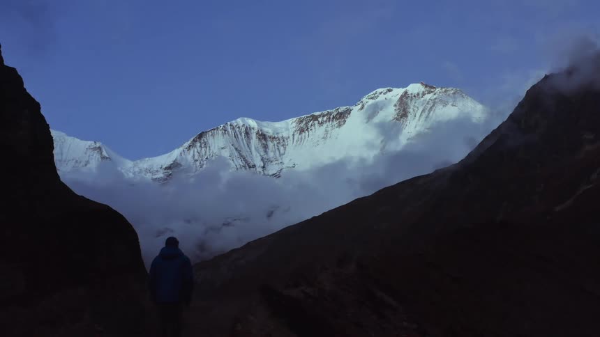 A hiker stands atop a mountain ridge while gazing at higher snow covered ridge in the distance