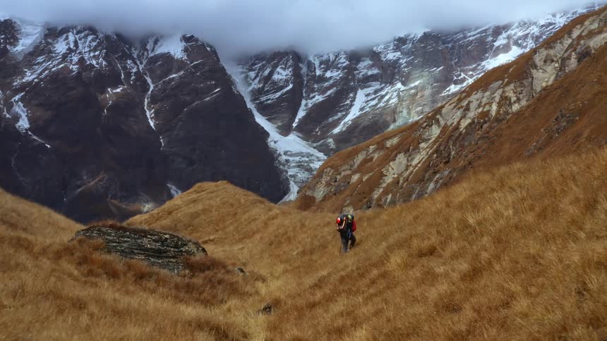 A pair of hikers walk through grassy hills at the base of a snowy mountain range