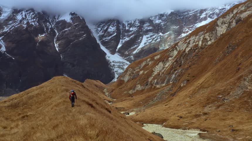 A hiker hikes along a hill covered in brown grass towards a snow capped mountain ridge