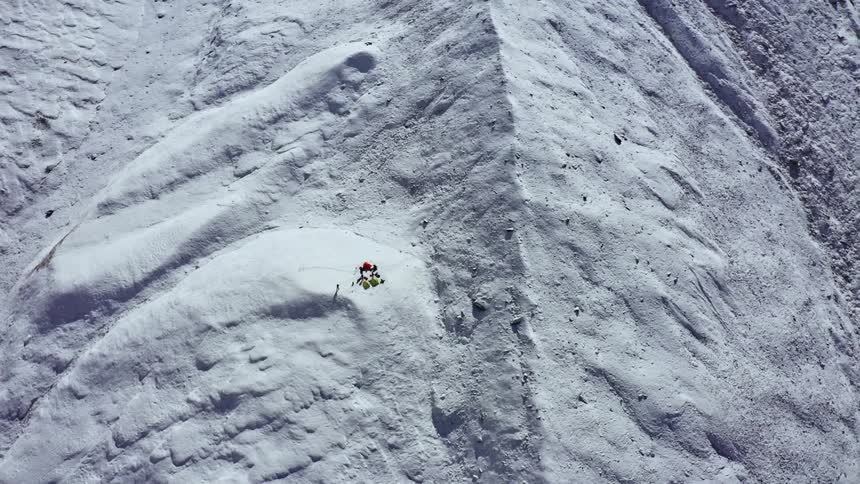 A group of people stand around a camp atop compacted snow on a mountain slope during the day