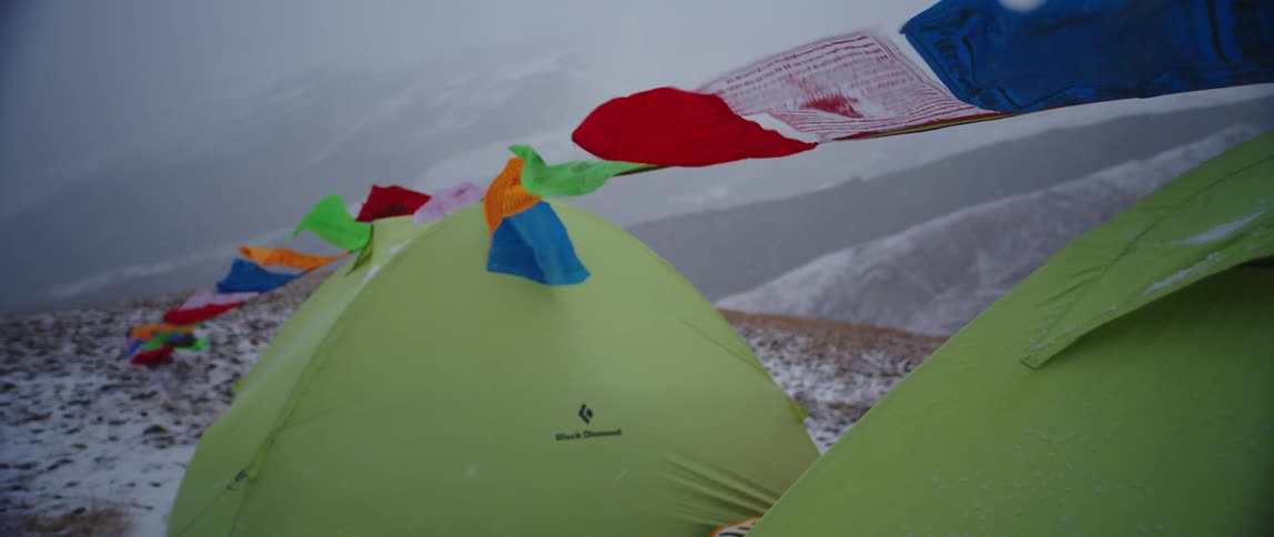Flags flap on rope tied among tents on snowy field with fog covered snowy mountain on horizon