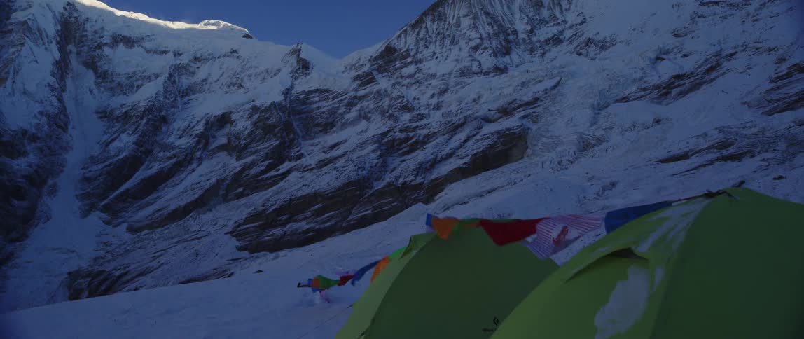 Banners flap on line near closed tents at base of snow covered mountain under clear blue sky