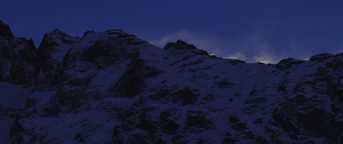 Thin clouds of snow drift above a snowy mountain ridge in the evening
