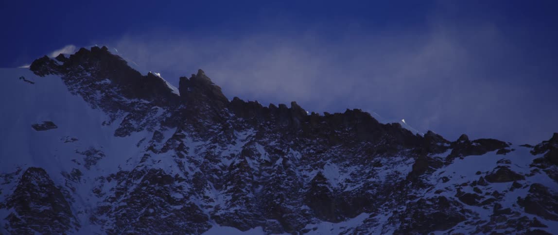 Snow blows over the summit of a mountain range in the evening