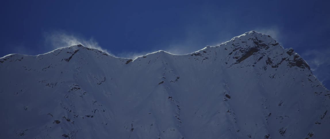 The wind blows clouds of snow off the summit of a mountain ridge on a sunny day