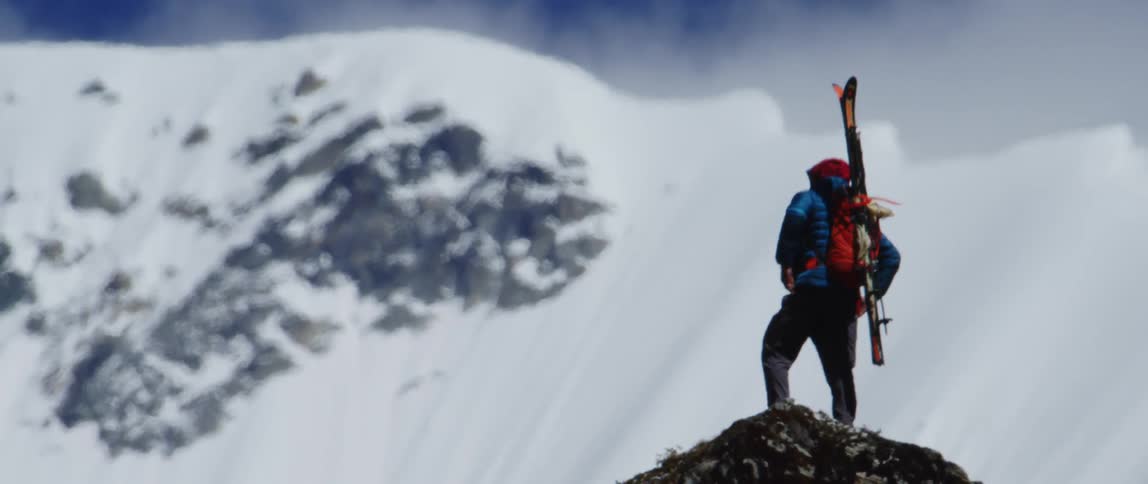 A hiker looks out over a snowy mountain range while standing atop a peak on a sunny day