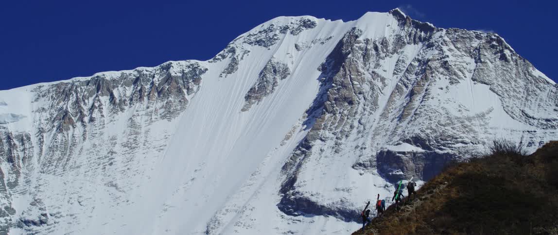 Four people with skis on their backs climbing up a hill with a mountain range in the background