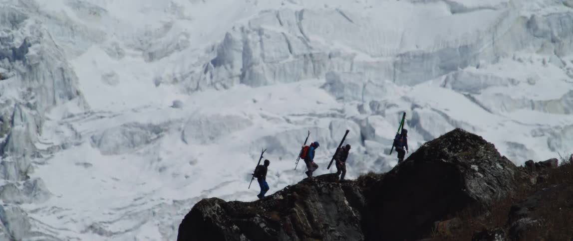 Four men with skis on their back hike up a mountain