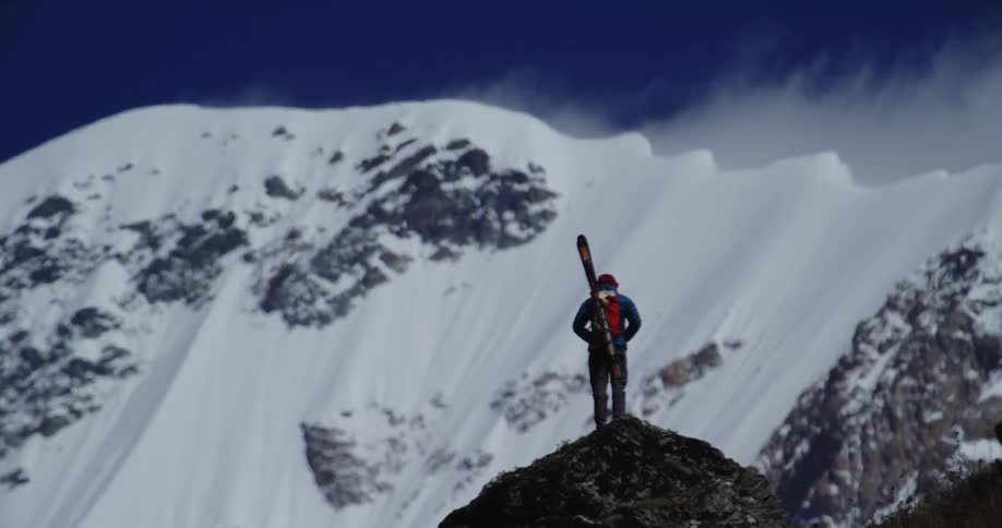 A hiker with skis strapped to their back looks out over a mountain range while standing on one of its peaks