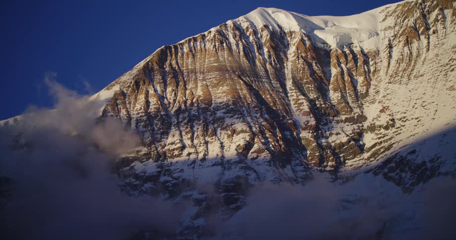 Clouds of fog float around a snowy mountain ridge on a sunny day