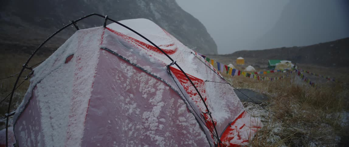 A tent in a grassy field covered in snow with a mountain range and the sky in the background