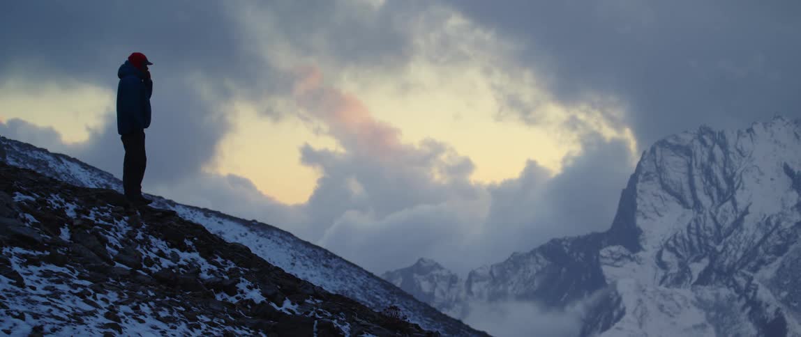 Man with a cap and a coat standing on a mountain slope watches snowy mountains