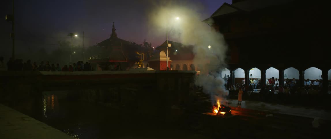 A fire burns beside a large pavilion and body of water at night