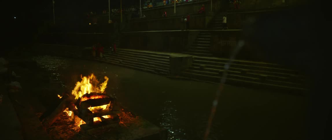 People walk along tiers of walkways near building across water from fountain and burning bonfire