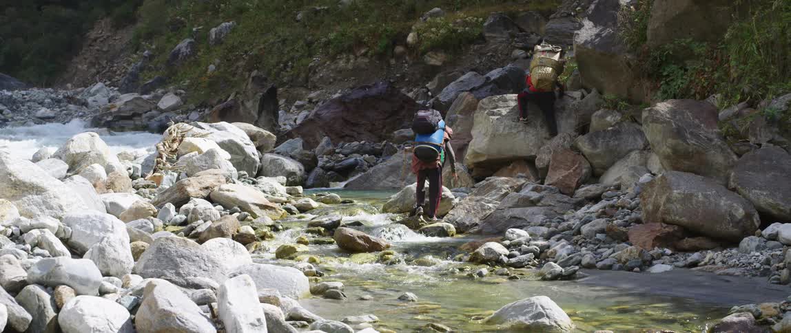 A hiker attempts to cross a stream atop a series of stones spanning it as another hiker attempts to climb a boulder to reach a slope