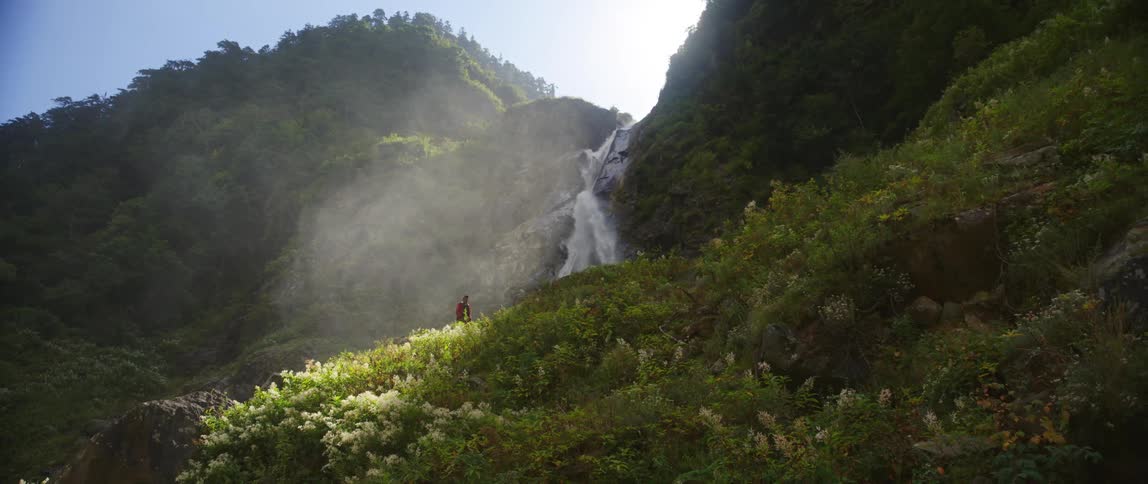 A person walks through a verdant mountain below a waterfall on a sunny day