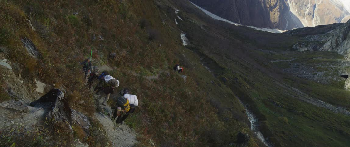 People carry packs along mountain trail with snowy covered pass in background
