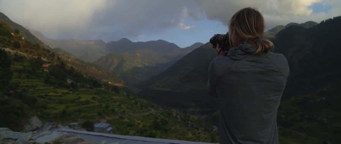 A photographer standing outdoors under a cloudy sky uses a camera to photograph a verdant mountain valley