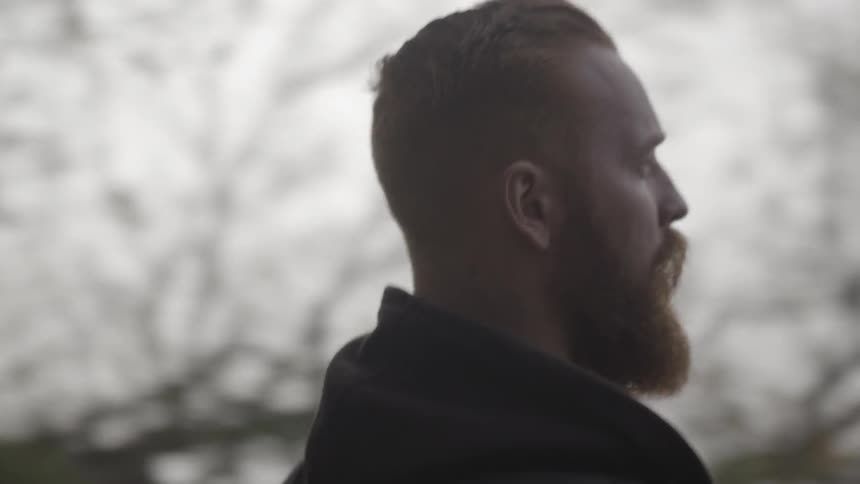 A bearded man walks in a forest toward a body of water