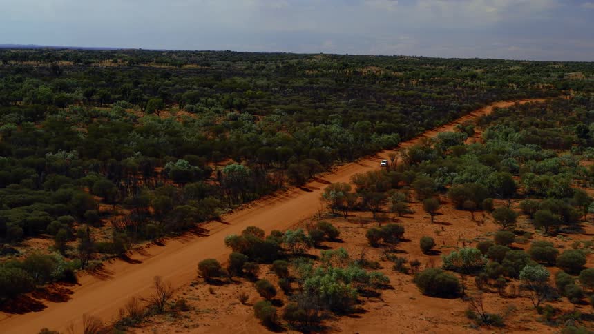 Aerial view of a white car driving on a red dirt road in the field