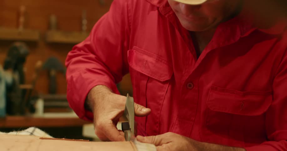 A man sits at a table and cuts excess fabric from a board piece