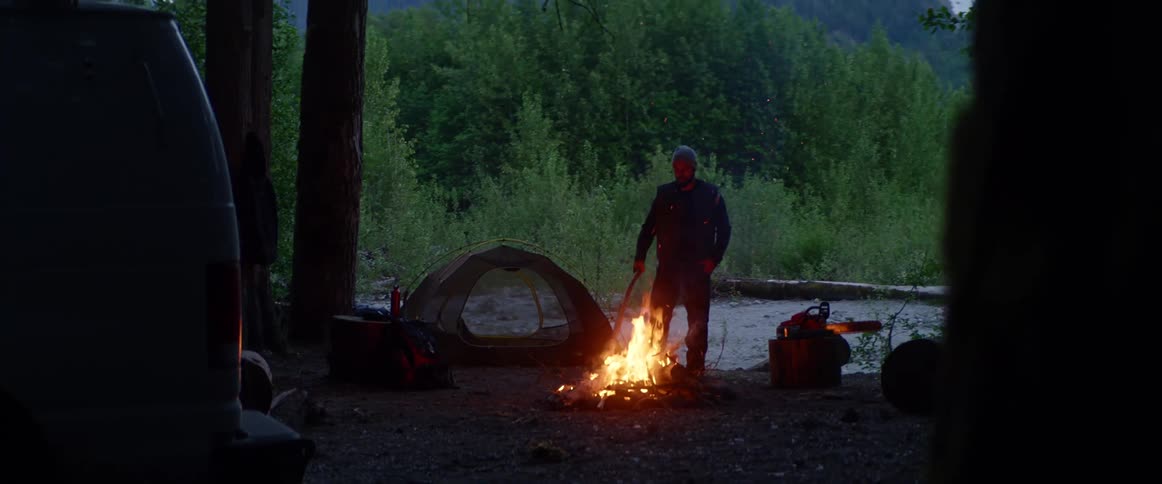 Man with a beard and a beanie stands between a tent and a campfire in the forest