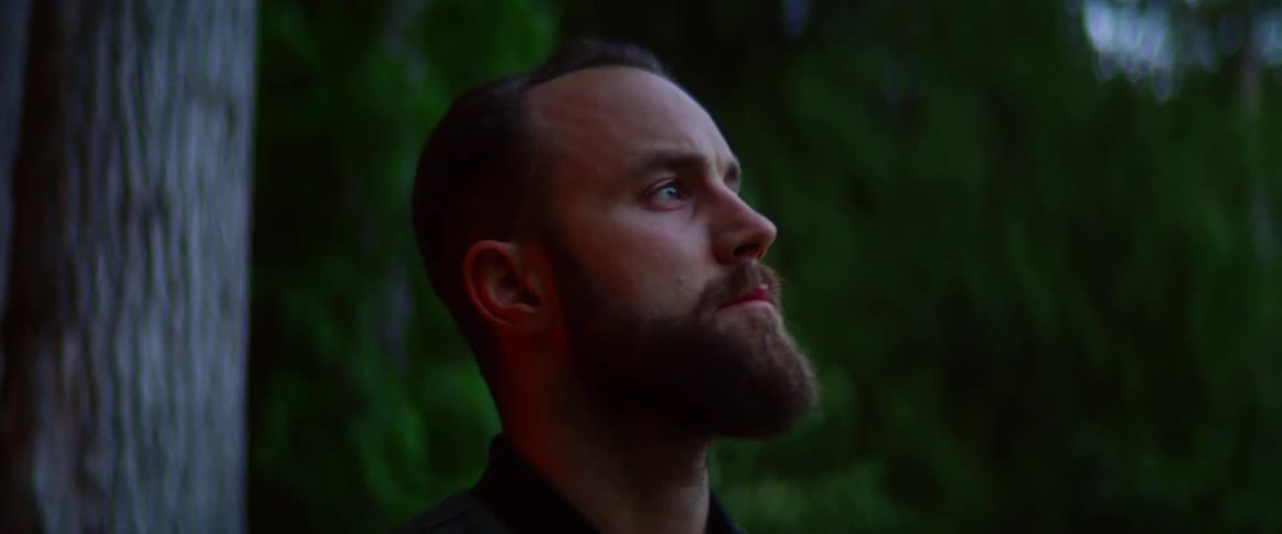 Side view of a man with a beard looking around in the woods at dusk