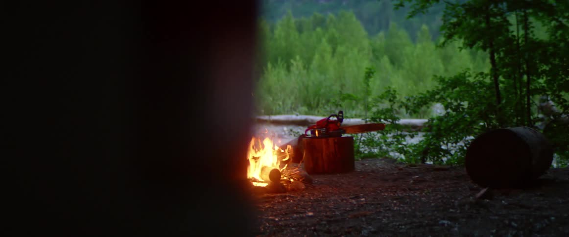 A man looks out over a verdant mountain valley while standing in front of his campfire and tent