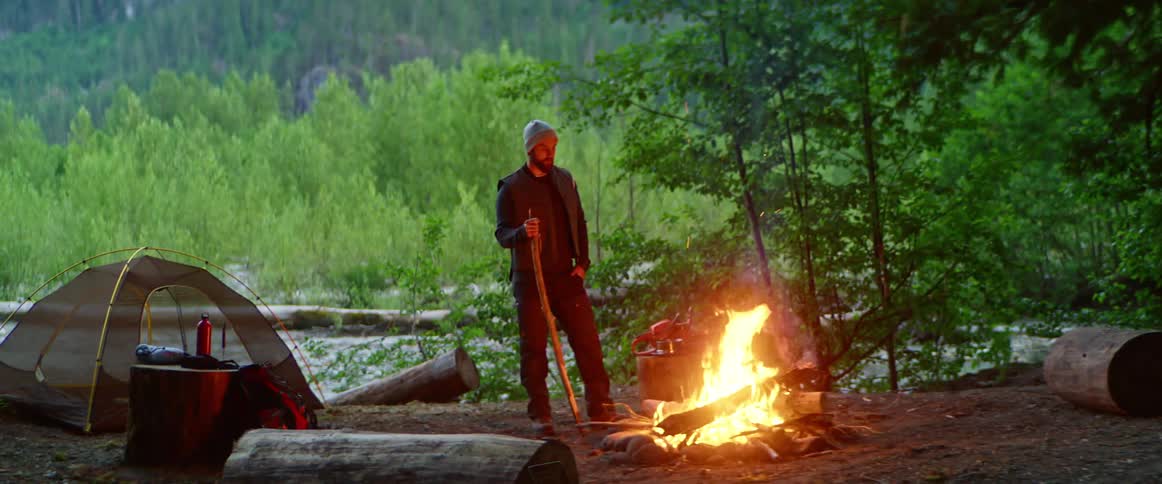 Man stands next to a campfire near his tent in the forest