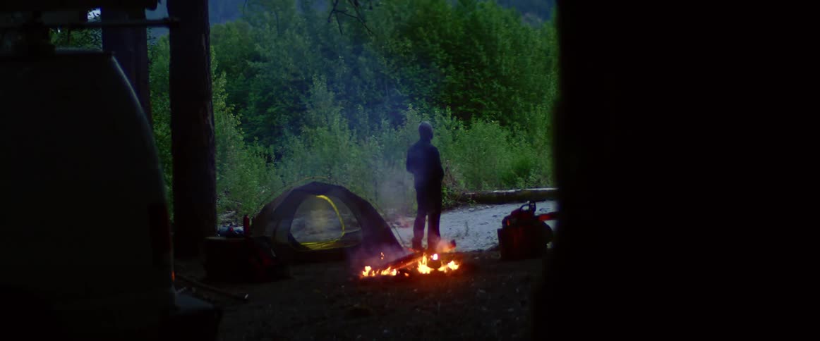 Man with a beanie and a coat standing next to a campfire is silhouetted against the trees in the forest