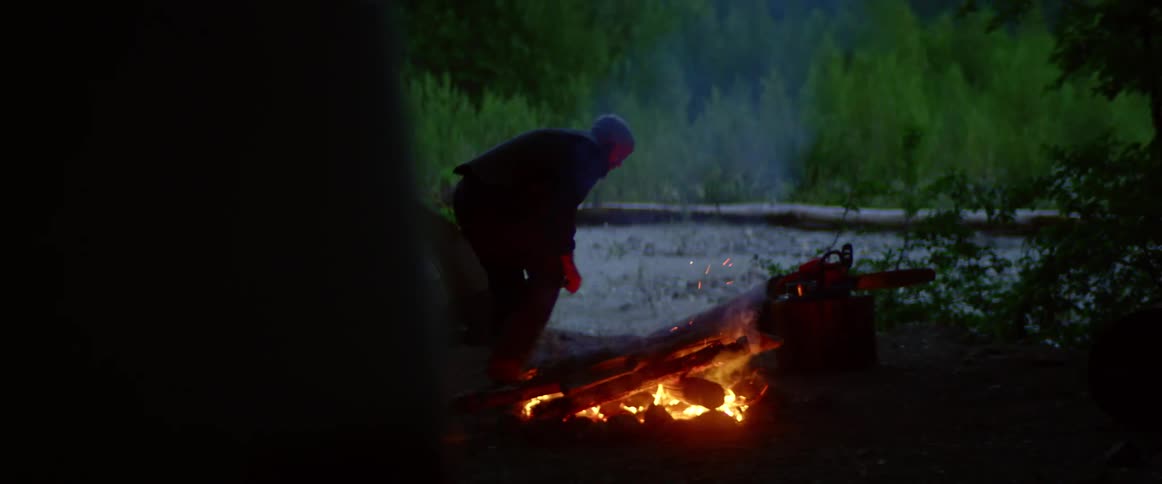 A man puts wood onto a campfire in a rural setting beside a small tent
