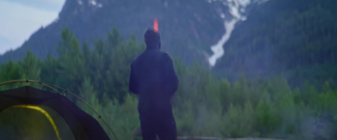 A man looks up at a verdant mountain from his campsite at its base as sparks from the fire float in the air