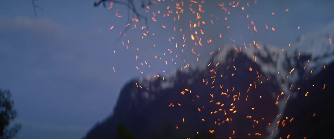 Branch turns in fire setting off sparks against tree branches and snow covered mountain