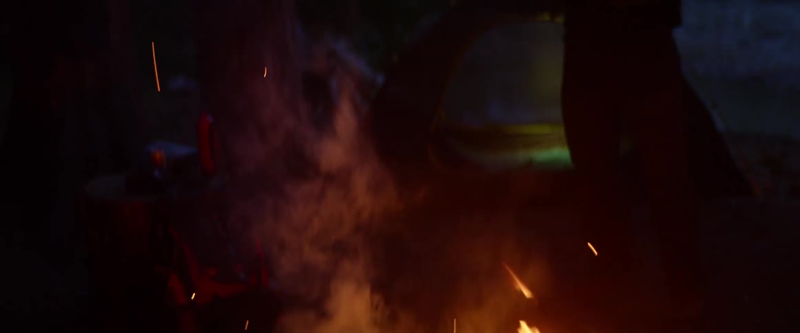Man with a beanie and a beard stands and watches a campfire in the forest at night