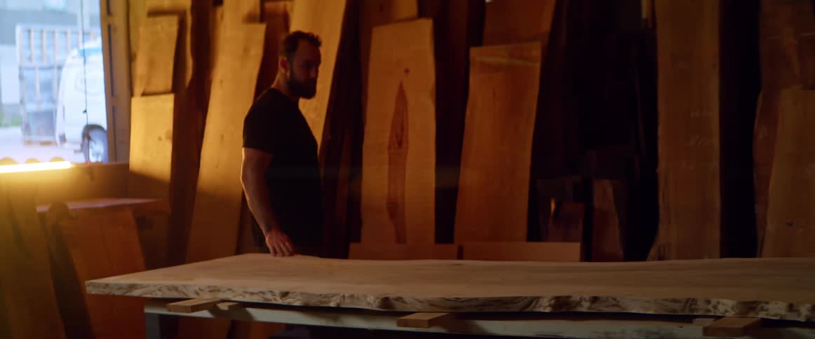A man runs his hand along a large slab of wood as he walks across a carpentry workshop