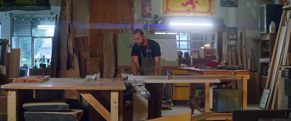 A man is cutting a piece of lumber on a table saw inside a workshop during the day