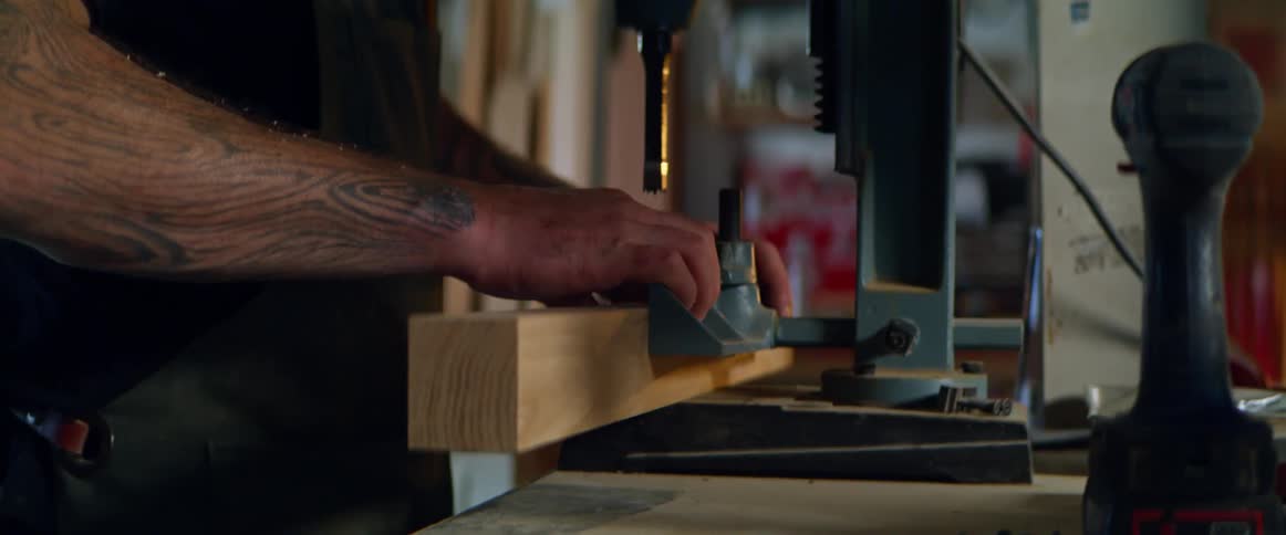 A man attempts to secure a block of wood under a drill press before giving up and sitting the block aside