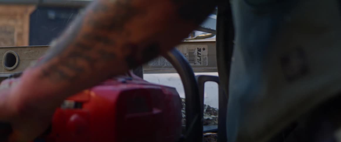 A tattooed man uses a chainsaw to slice a log outside of a building during the day